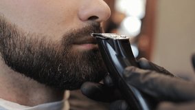 Barber shaving bearded man in barbershop. Male skin care and beard style concept. Master hairdresser with a professional trimmer in a black gloves makes a haircut and evens the beard to a customer.