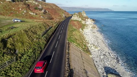 Northern Ireland, UK, Atlantic coast with cliffs. Red car on Antrim Coast Road a.k.a. Causeway Coastal Route. One of the most scenic coastal roads in Europe. Aerial 4K video. Winter, sunrise light