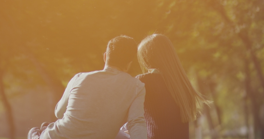 Back view of happy lovely caucasian romantic couple sitting on lawn and hugging each other in park . Rear view of young couple sit on grass and enjoy in the open air. Happiness people . Slow Motion .