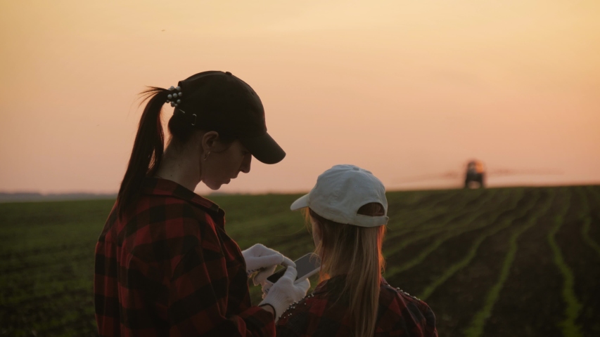 Family farmers work in the field, look at the digital tablet against the background of the tractor. The farmer mother and her daughter talk in the field, use a tablet. Smart and modern farm concept.