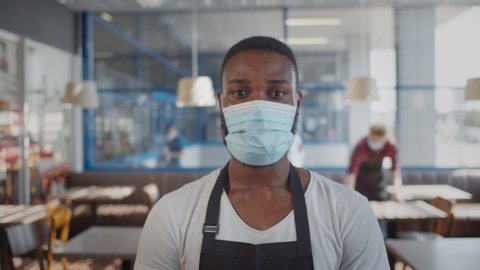 Portrait of afro-american waiter wearing protective mask looking at camera over blurred background of cafe interior. Young african coffee shop owner or barista in apron and safety mask