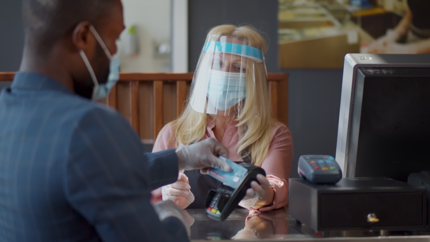 Back view of afro-american customer in protective mask and gloves paying for takeaway order with contactless credit card in cafe. Mature cashier in plastic face shield with card reader at counter | Shutterstock HD Video #1056077567