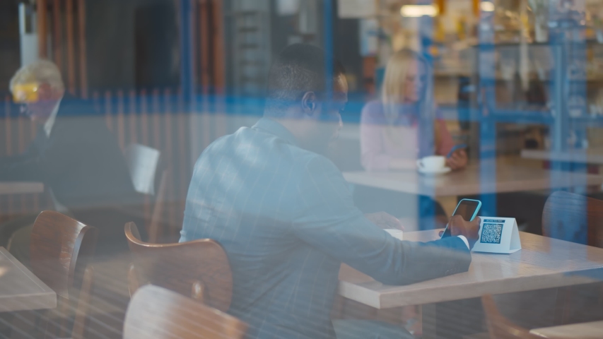 Handsome afro businessman scan barcode in restaurant. View through window of young afro-american male customer sitting at table and scanning qr-code with mobile phone for contactless payment in cafe. - Powered by Shutterstock - Get 15% off with code: PIKWIZARD15