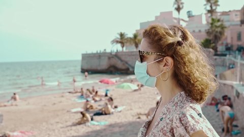 Beautiful curly brunette tourist with a surgical mask during Covid-19 with the Mediterranean village of Sitges on the bakground. Safe Travel in the new normal.