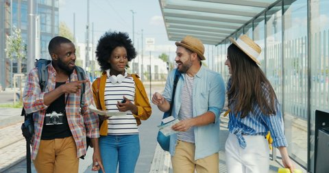 Multiethnic males and females tourists walking together at bus stop and talking cheerfully on summer day. Group of mixed-races stylish happy men and women travellers strolling and having fun.