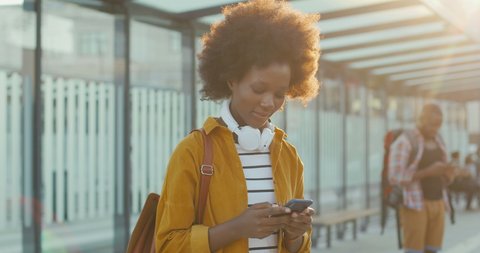 Young African American woman texting message on smartphone and smiling cheerfully at train station. 