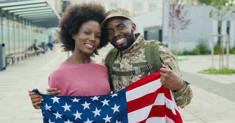 Portrait of joyful African American couple hugging and smiling outdoors at street. Handsome happy man soldier in military uniform hugs and embracing girlfriend with USA flag. Meeting of guy and girl.