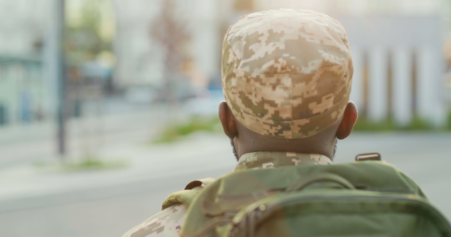 Rear of African American young male soldier in uniform, hat and with backpack walking outdoor. Back view on militarian officer strolling the street in military clothes. Going to army. Royalty-Free Stock Footage #1056080669