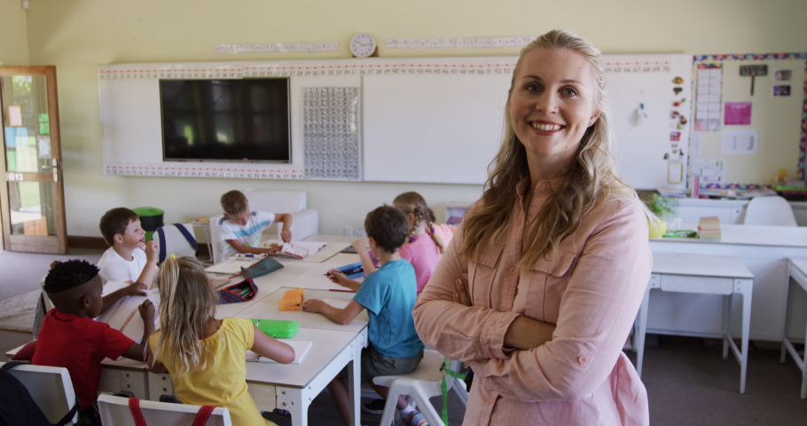 Caucasian female teacher standing in a classroom with arms crossed, looking at camera and smiling, with her pupils sitting at desks in the background, in slow motion. education at an elementary school | Shutterstock HD Video #1056082196