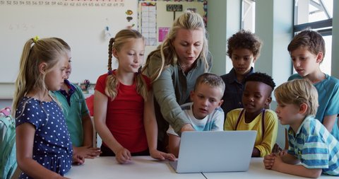 Multi-ethnic group of children and their Caucasian female teacher sitting in a classroom during lesson, using a laptop computer, in slow motion. Education at an elementary school.