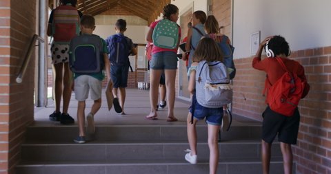 Multi-ethnic group of children wearing backpacks, walking in an outdoor corridor during break, in slow motion. Education at an elementary school.