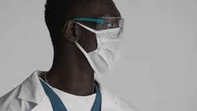 Close up young African American male doctor in protective eyeglasses and medical mask confidently looking in camera isolated