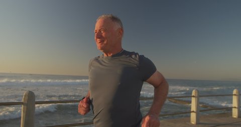 Senior fit Caucasian man working out on promenade by the sea wearing sports clothes, running on a sunny day in slow motion. Retirement healthy lifestyle activity.