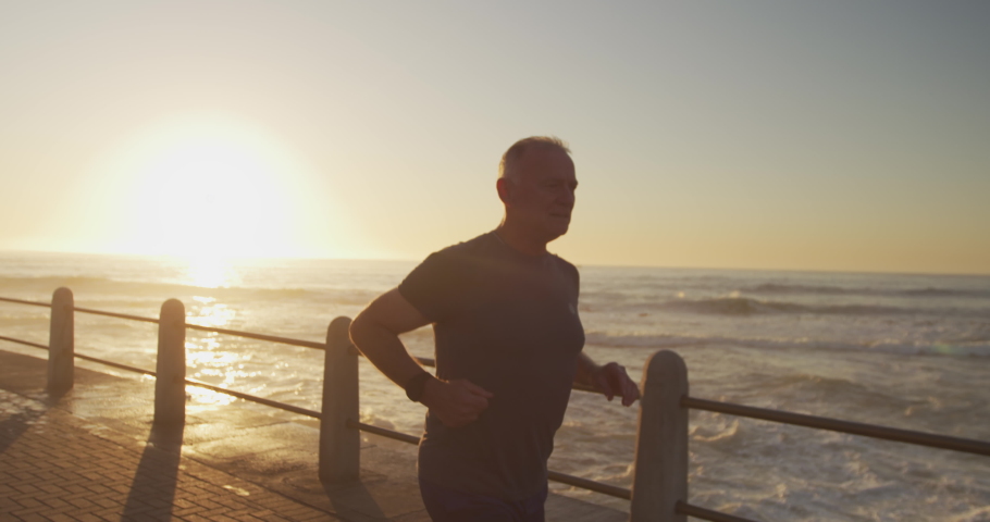 Senior fit Caucasian man working out on promenade by the sea wearing sports clothes, running on a sunny day in slow motion. Retirement healthy lifestyle activity.