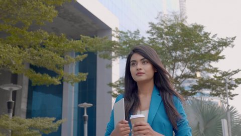 A confident corporate office woman employee in formal dress holding a coffee mug and laptop in hands walking along the street. An attractive and beautiful businesswoman outside the commercial complex.