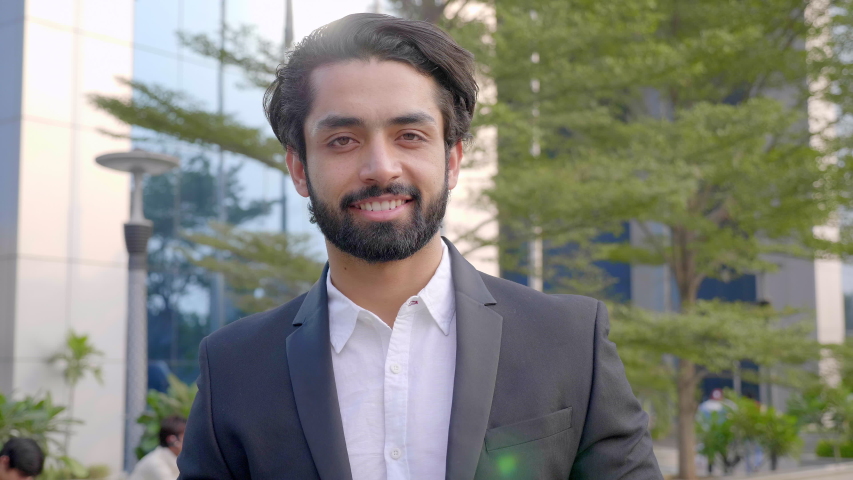 A well dressed happy bearded office corporate male executive standing outside a modern corporate building. A handsome businessman smiling and looking into the camera outside modern commercial complex | Shutterstock HD Video #1056089255