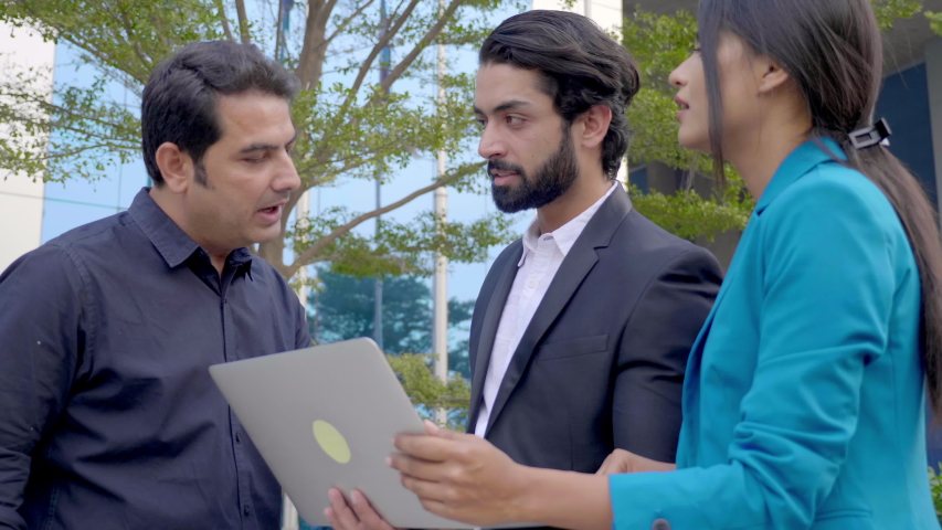 A team of business partners standing outdoors and discussing an upcoming project using a laptop. A group of male and female corporate office employees interacting outside the commercial complex  Royalty-Free Stock Footage #1056089258