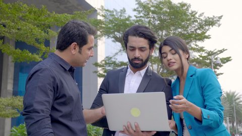 A team of business partners standing outdoors and discussing an upcoming project using a laptop. A group of male and female corporate office employees interacting outside the commercial complex 