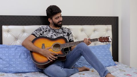 A bearded Indian man practicing his new lyrical song on guitar - music concept. Portrait of a handsome young musician in wired earphones, enjoying singing while playing guitar - leisure time