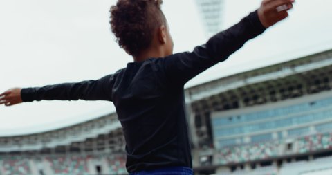Portrait of cute little black kid boy spreading hands on a large football stadium, dreaming of becoming professional player, soccer star. Shot on RED camera with anamorphic lens