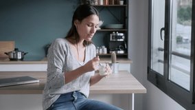 Video of smiling young woman eating yogurt while sitting on stool in the kitchen at home.