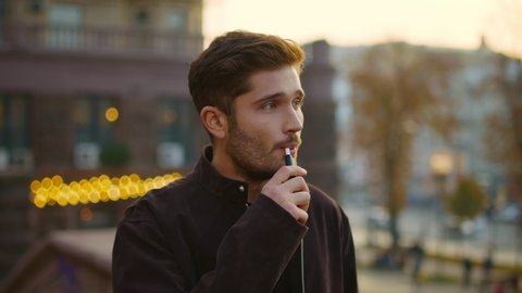 Portrait of serious hipster man smoking electronic cigarette on city street. Closeup thoughtful guy walking with e-cigarette in hand. Handsome male person vaping in urban background.