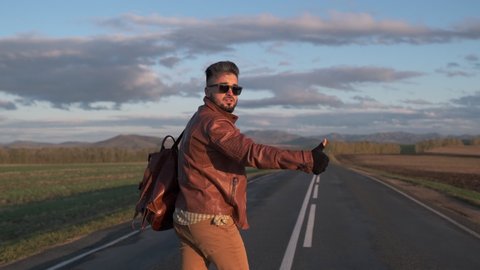 Hipster hitchhiker tourist traveler man with backpack walking along long road, hitchhiking lifting thumb up in summer to stop car. trip hitching mountain nature travel tourism adventure, 4 K slow-mo