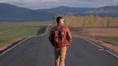 Hipster hitchhiker tourist traveler man walking along long road, hitchhiking summer. backpack backpacking traveling trip hitching mountain nature vacation travel tourism freedom rear view, 4 K slow-mo