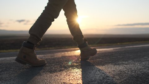Close up of feet boots of tourist traveler man walking along road, hitchhiking at sunset. backpacking traveling step way trip hitching nature vacation travel tourism freedom sun lens flare 4 K slow-mo