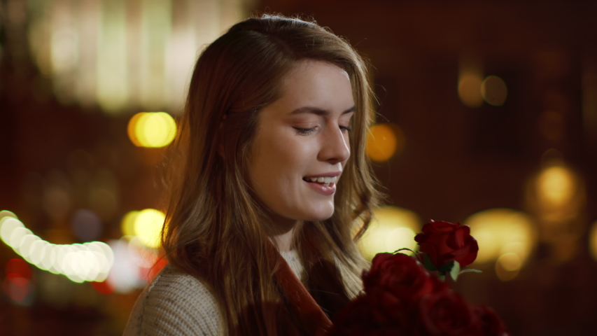 Closeup romantic woman smelling bouquet of flowers on night street. Portrait of affectionate girl holding red roses in hands outdoors. Gorgeous woman enjoying floral bunch in urban background.