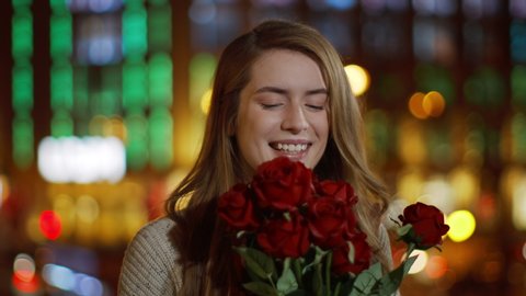 Closeup romantic woman smelling bouquet of flowers on night street. Portrait of affectionate girl holding red roses in hands outdoors. Gorgeous woman enjoying floral bunch in urban background.