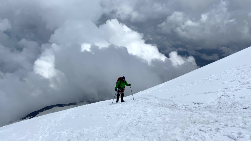 A climber with a backpack and trekking poles walks along a snowy path to Elbrus. Aerial view of a beautiful mountain winter landscape. Snowy rocky slopes of the northern Elbrus region.