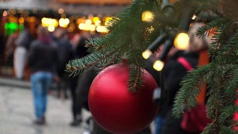 Closeup of a red Christmas ball hang on a natural New Year tree on a city fair, crowd of people walking on a festive street.
