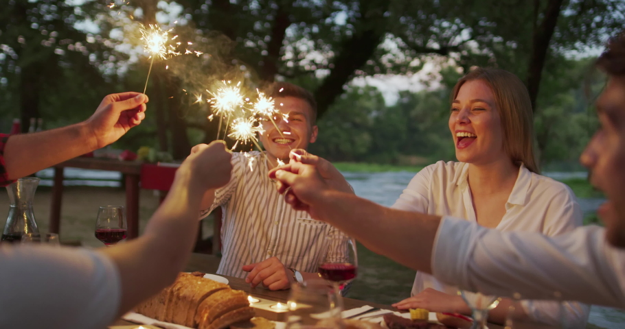 Group of happy friends celebrating holiday vacation using sprinklers and drinking red wine while having picnic french dinner party outdoor near the river | Shutterstock HD Video #1056128312