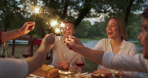 Group of happy friends celebrating holiday vacation using sprinklers and drinking red wine while having picnic french dinner party outdoor near the river