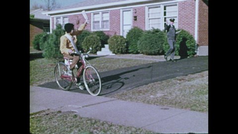 1970s: Boy kneels on lawn, loads newspapers into bag, picks up bag, gets on bicycle and rides away. Boy rides bicycle down street, throws newspapers to houses, waves at mailman.