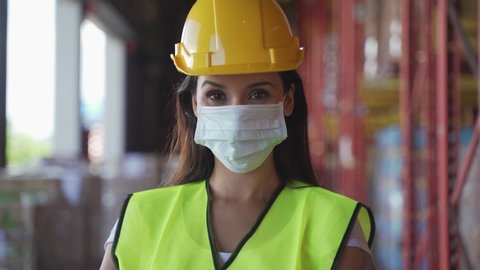 Portrait female worker with safety vest, hard helmet wearing face mask standing in warehouse factory due to covid pandemic crisis. Girl working in logistic and storage industrial with care of health.