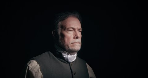A mature bearded man 18th century American colonist type in a dark room whose face is lit by a sole light source turns and looks solemnly into the camera.