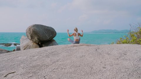 Pretty young slim woman meditating in lotus pose on the rocks by the tropical ocean beach, doing yoga on top of the mountain on a rock in nature. Healthcare, wellness, mindfulness concept.