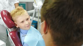 Dentistry concept. Child patient sitting on dental chair in paediatric dentists office. Assistant putting and fixing napkin. Boy patient at dental clinic getting treatment. 4 k video