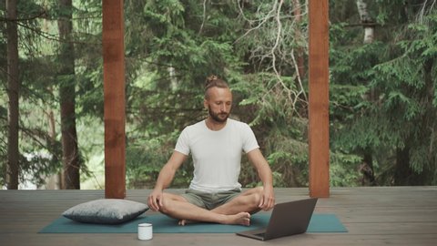 Handsome office man in white t-shirt try to meditate with online guiding on laptop. Beginner in yoga practice checks when timer ends. Difficult to sit long time. Terrace on green forest background 