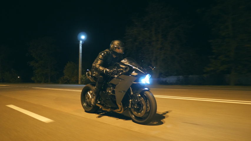 Young man in helmet riding fast on modern black sport motorbike at evening city street. Motorcyclist racing his motorcycle on night empty road. Guy driving bike. Concept of freedom and hobby. Close up