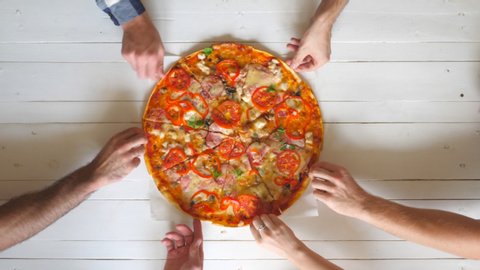 Top view male and female hands taking slices of pizza with cheese, tomatoes and ham from food delivery. Group of hungry friends sitting at desk and sharing delicious lunch on wooden table background