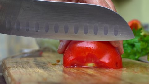 Female cook cuts ripe red tomato on a wooden cutting board.
Chopping sliced tomato into small pieces with a kitchen knife.
Concept of homemade food and cooking.
Close up. Selective focus.
