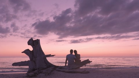 Romantic beach sunset couple honeymoon - Lovers enjoying watching sunset on summer travel destination sitting on tree trunk by the ocean. From Lovers key, Florida near Fort Myers Beach, USA.