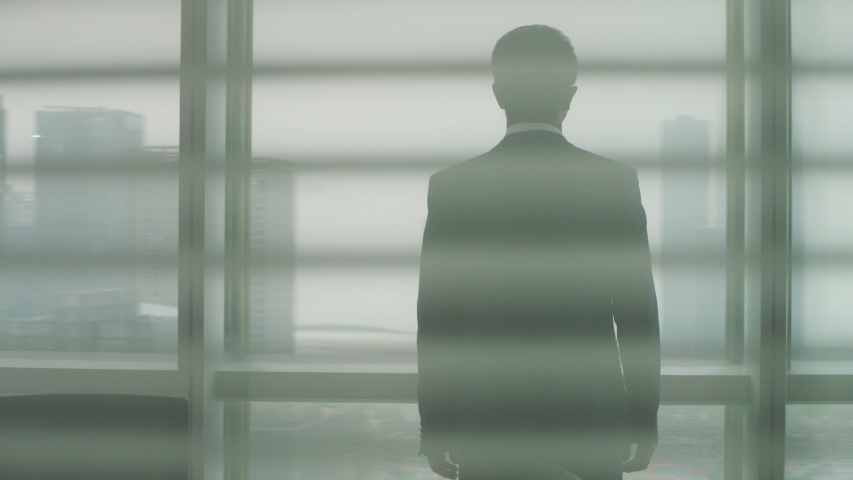 rear view of a mature asian business man standing by the window in office looking out at modern buildings  Royalty-Free Stock Footage #1056144245