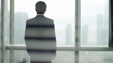 rear view of a mature asian business man standing by the window in office looking out at modern buildings 