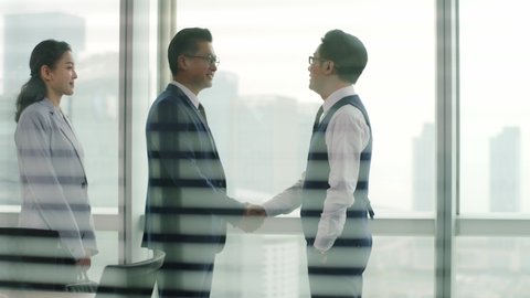 young asian corporate executive welcoming and shaking hands with visiting client in front of windows in office