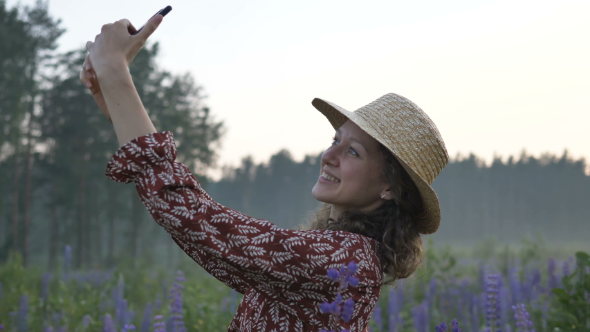 happy young lady wearing stylish straw hat and dress with floral pattern makes selfie with phone among lupin flower field slow motion