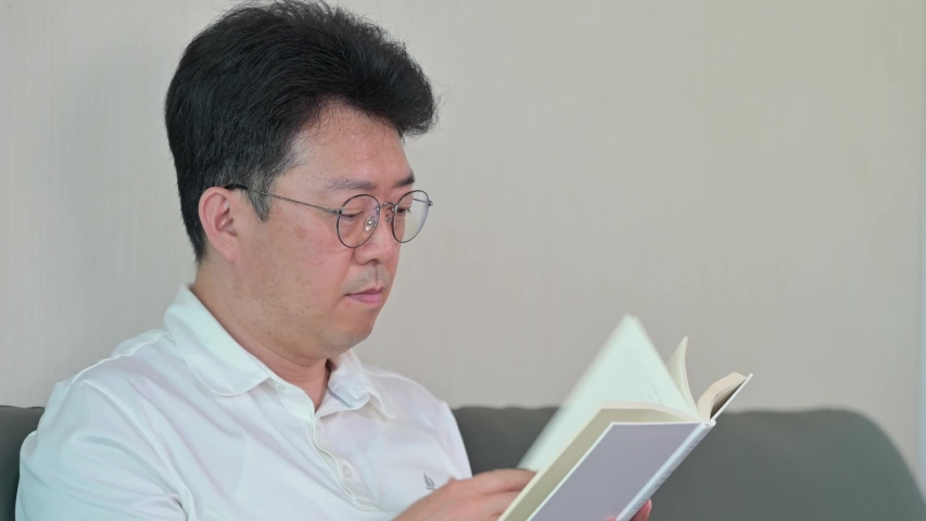 An Asian middle-aged man reading in the living room with an air purifier on. Zoom Out.